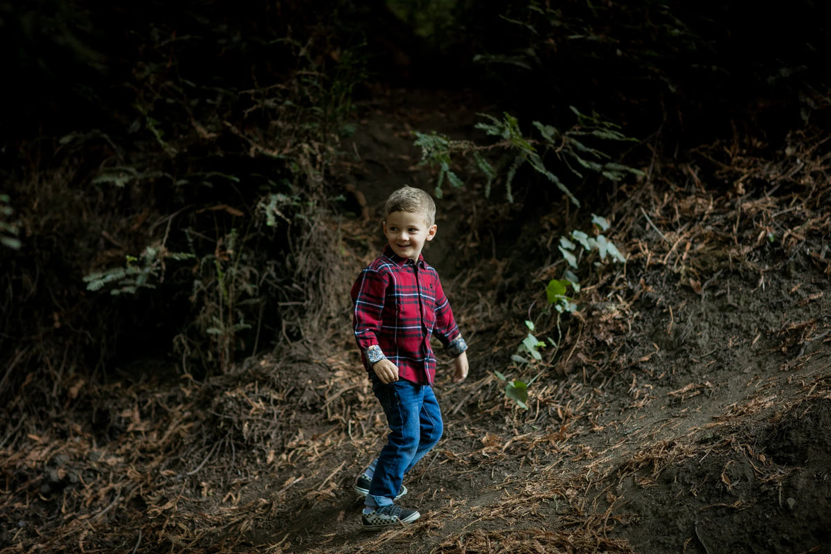 A young boy in a red plaid shirt walks on a forest path, smiling and surrounded by greenery and fallen leaves.