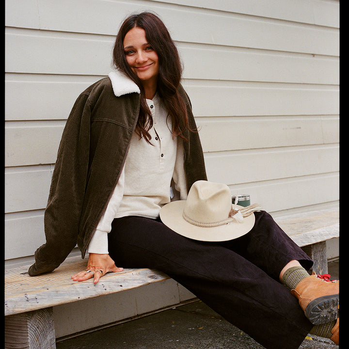 A woman with long brown hair sits on a wooden bench against a light wall, smiling. She wears a Women&