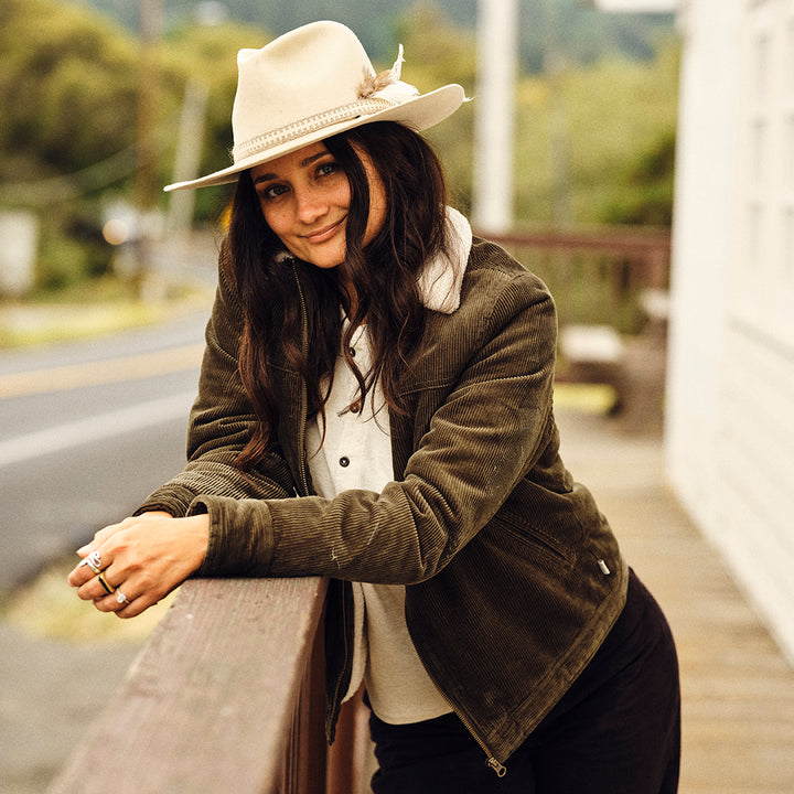 A woman with long dark hair smiles outdoors, leaning on a wooden railing. She wears rings, a light wide-brimmed hat, and the Women&