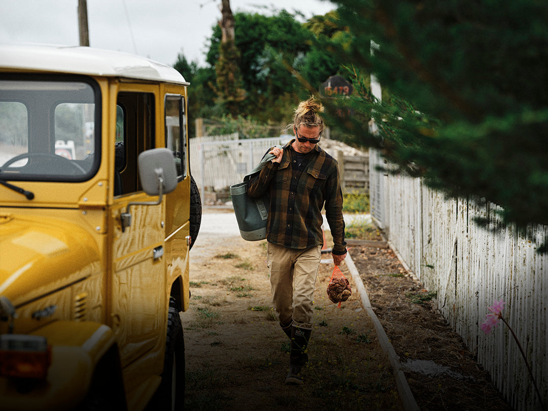 A person walking outdoors beside a yellow vehicle and wearing a plaid shirt, carrying a dark tote bag.