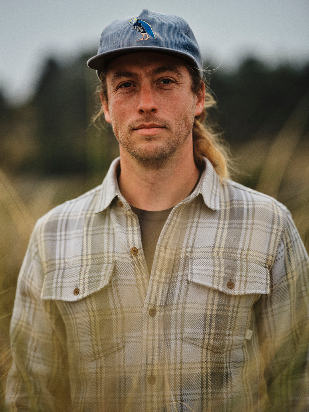 Man with long hair in a plaid shirt and blue cap standing outdoors in a grassy field, blurred trees in background.