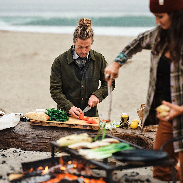 Two people cook by the beach; one in a Pladra Men's Sequoia Chore Coat—Hunter Green chops veggies while the other grills.