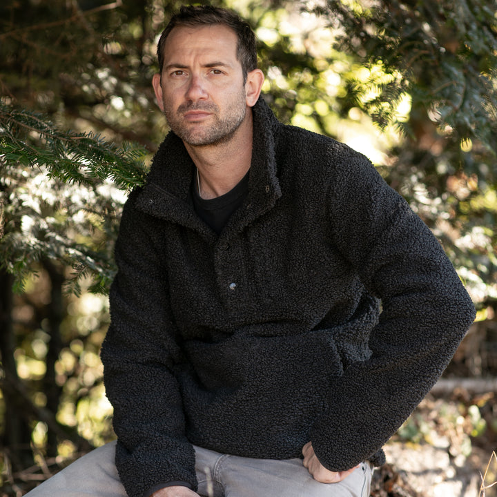A man with short dark hair and stubble sits outdoors among green trees, wearing the Men&