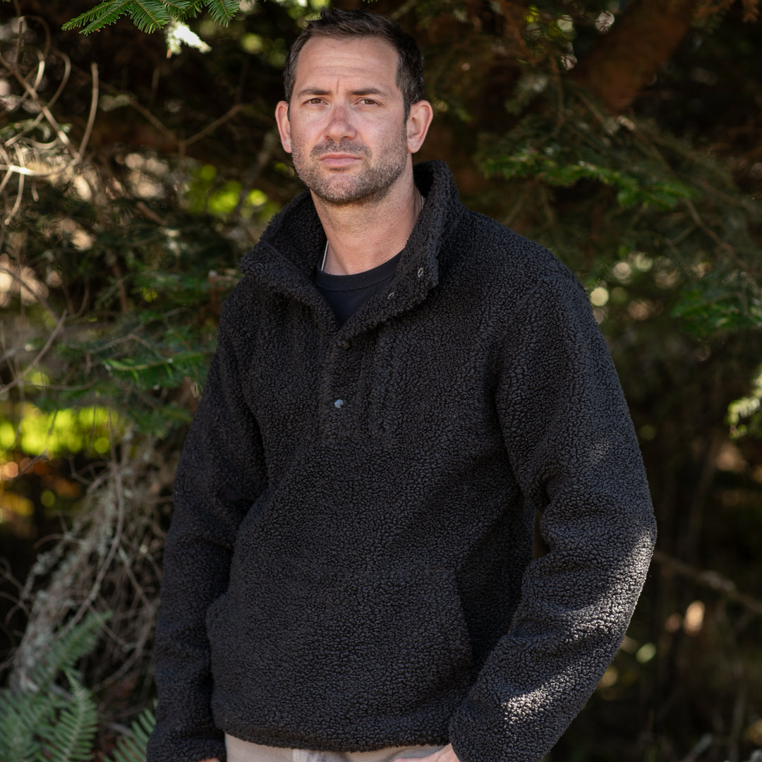 A man with short dark hair and a trimmed beard stands outdoors in front of sunlit green trees, wearing the Men&