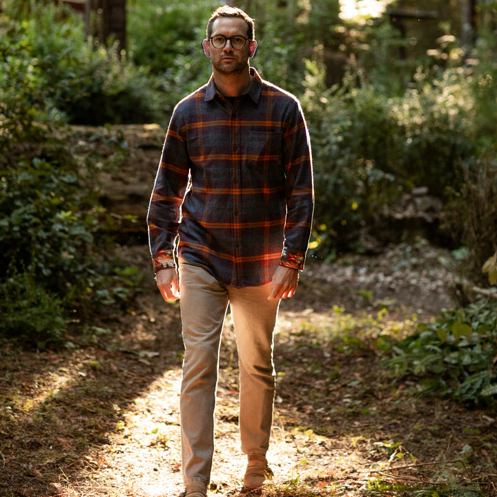 Man in Pladra Men’s Every Day Flannel Shirt—Portsmouth Blue Heather walks a sunlit forest path in khaki pants.