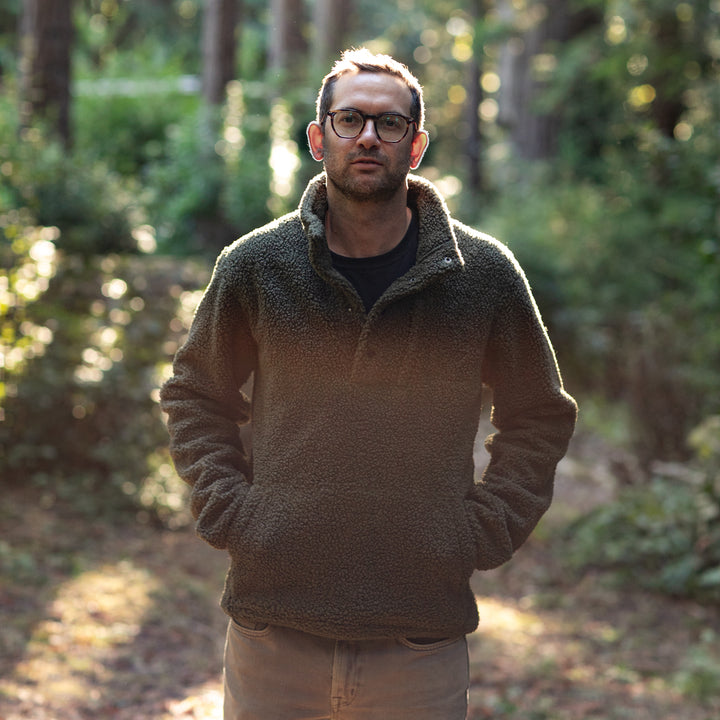 A man wearing glasses and the Men's Cypress Sherpa Fleece in Forest Green stands in a sunlit forest, hands in his pockets, surrounded by lush foliage and gentle natural light.