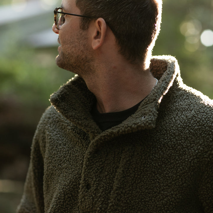 A man in glasses wears the Men's Cypress Sherpa Fleece in Forest Green, looking to his left outdoors with sunlight streaming through trees in the background.