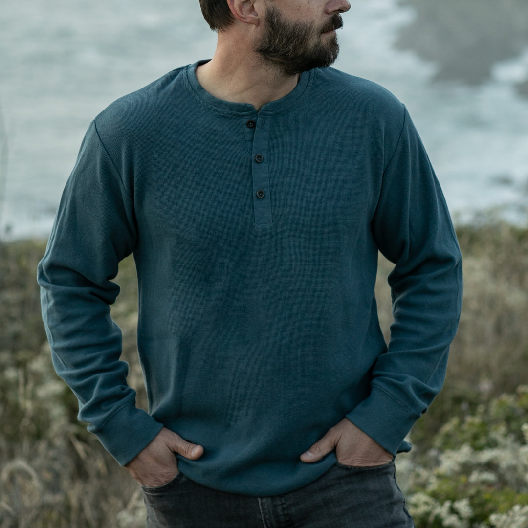 A bearded man stands outdoors with his hands in his pockets, wearing the Men’s Elko Henley in Atlantic Blue and dark jeans, with blurred greenery and water in the background.