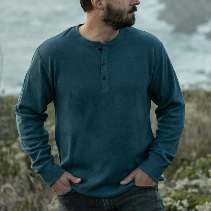 A bearded man stands outdoors with his hands in his pockets, wearing the Men’s Elko Henley in Atlantic Blue and dark jeans, with blurred greenery and water in the background.