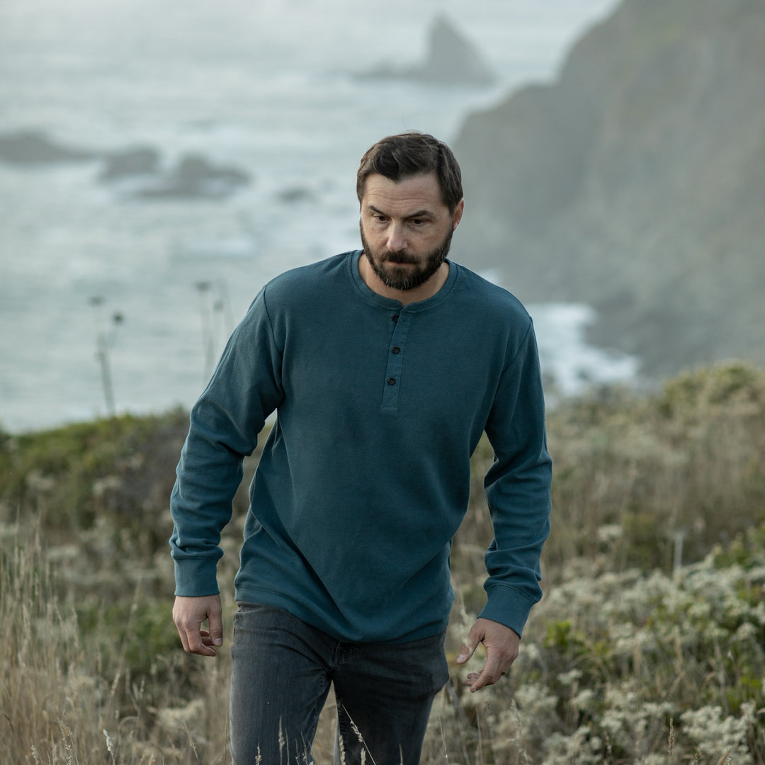 A bearded man wearing the Men's Elko Henley in Atlantic Blue and jeans walks through tall grass on a coastal cliffside, with the ocean and rocky shore behind him.