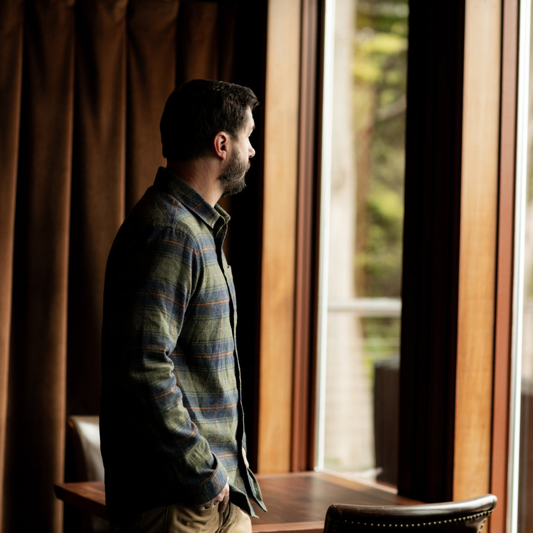 A man with dark hair and a beard stands indoors by a large window, wearing the Men&