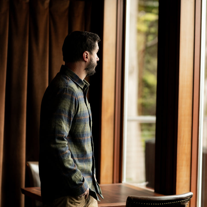 A man with dark hair and a beard stands indoors by a large window, wearing the Men&