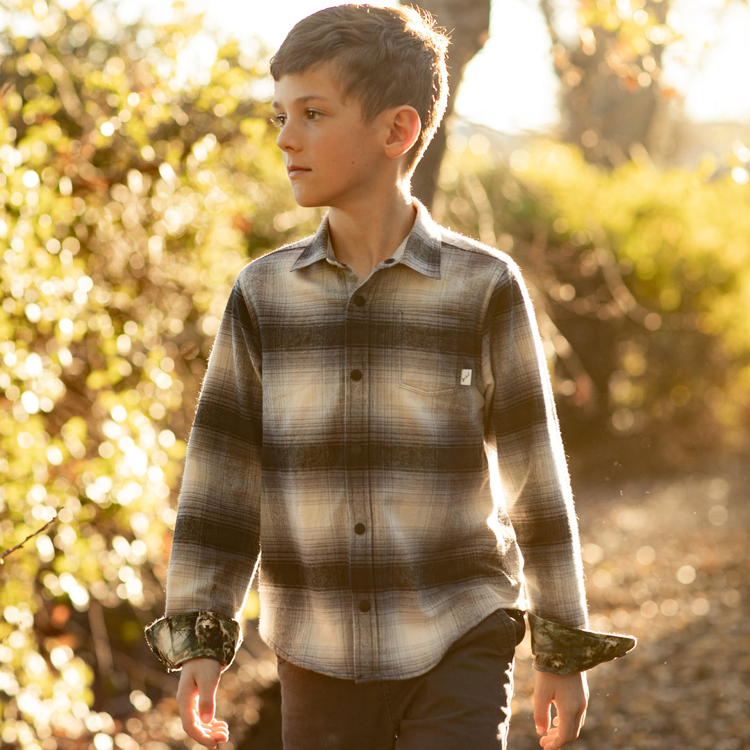 A boy in a Pladra Toddler Cubs Flannel Shirt—Sterling Grey Heather walks outside in sunny, green surroundings.