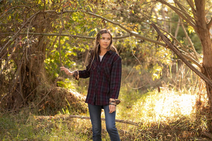 A young woman stands outdoors in a sunlit, wooded area, gently holding a tree branch and looking thoughtfully to the side while wearing the Women&