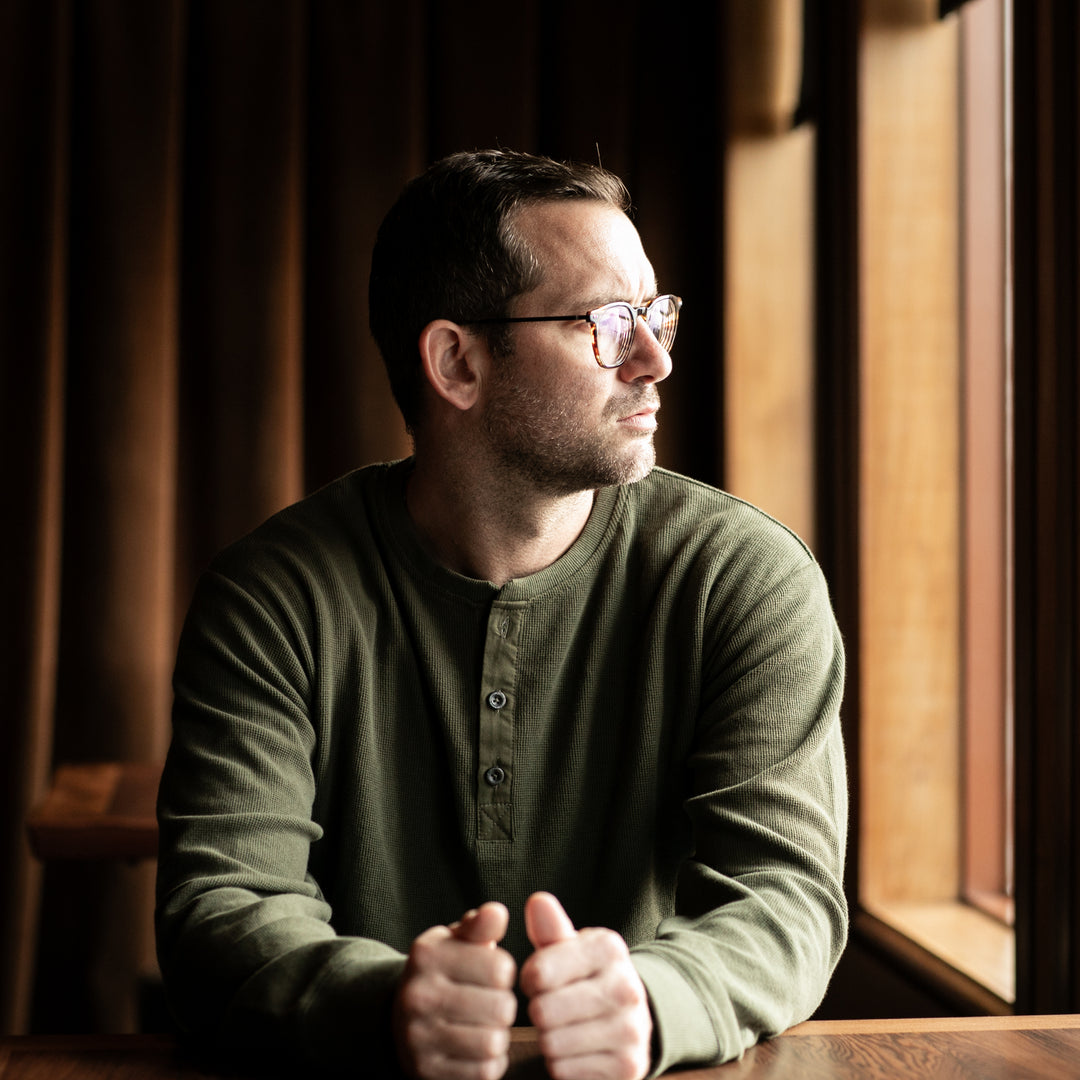 Man in glasses and forest green Pladra Men's Elko Henley sits thoughtfully by a sunlit window.