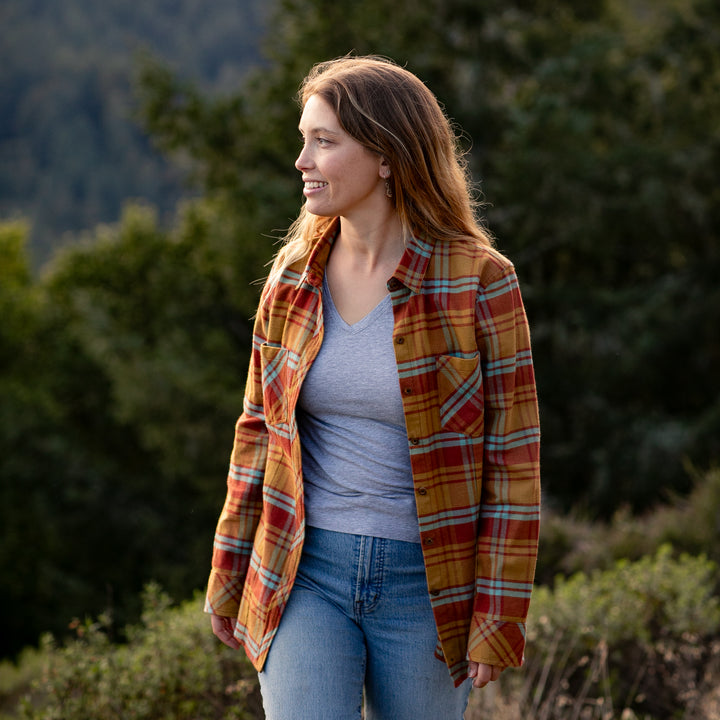 A woman stands outdoors among trees, smiling and looking to the side, wearing the Women’s Every Day Flannel Shirt–Cottonwood Orange over a gray tee and jeans. The premium cotton fabric adds comfort to her casual style.