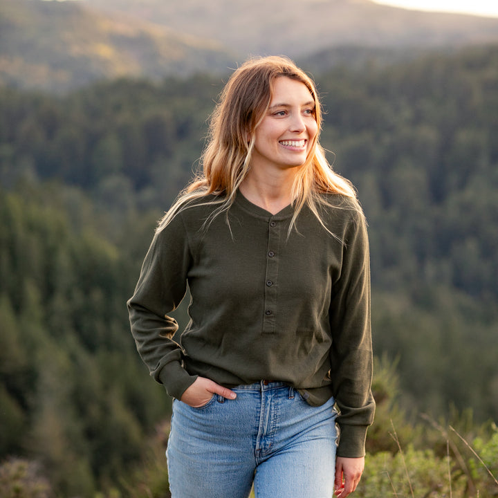 A young woman with long, wavy hair smiles outdoors, wearing the Women&