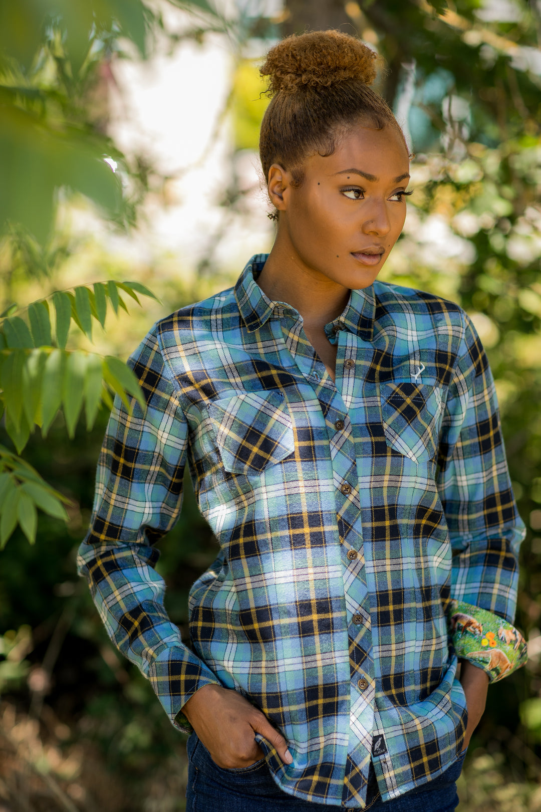 A woman in a blue plaid shirt stands outdoors among green trees, looking thoughtfully to the side.