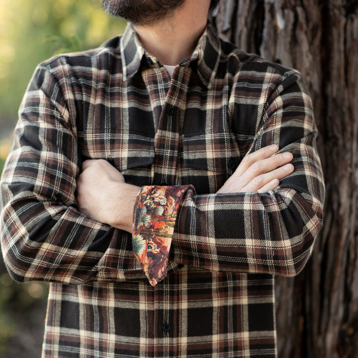 A bearded man stands outdoors with arms crossed, wearing the Men&