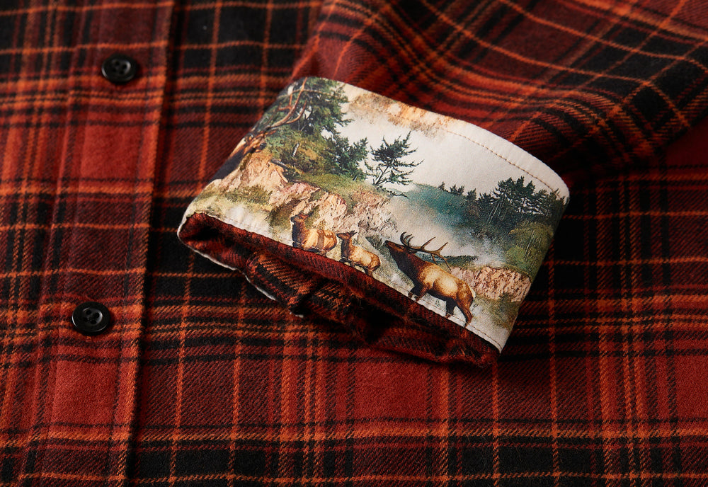 A close-up of the Men's Every Day Flannel Shirt- Autumn Red shows its red and black plaid pattern, with a rolled-up cuff revealing a printed inner lining that features an outdoor scene with trees and elk.