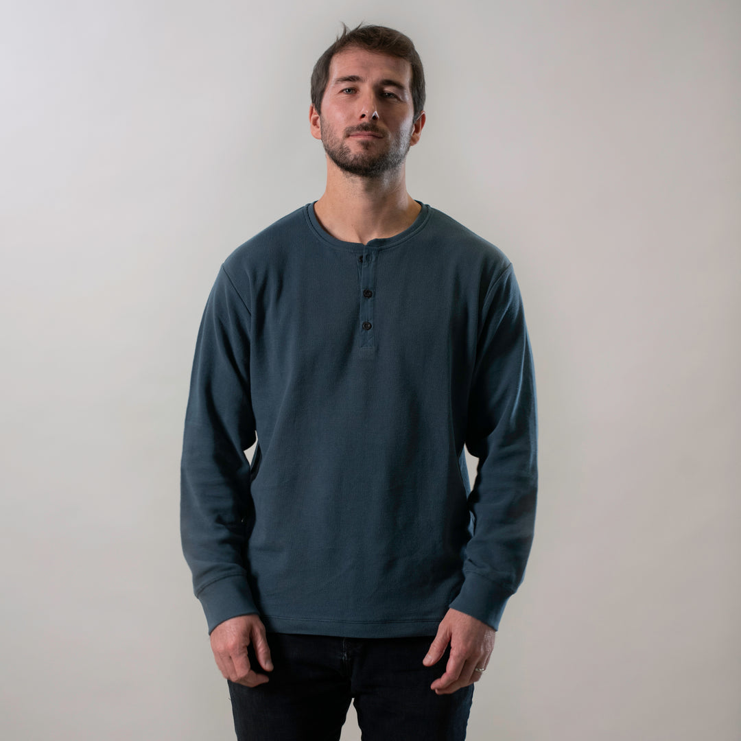 A man with short brown hair and a beard stands against a plain light background, wearing the Men's Elko Henley in Atlantic Blue and black pants, with a neutral expression and his hands relaxed by his sides.