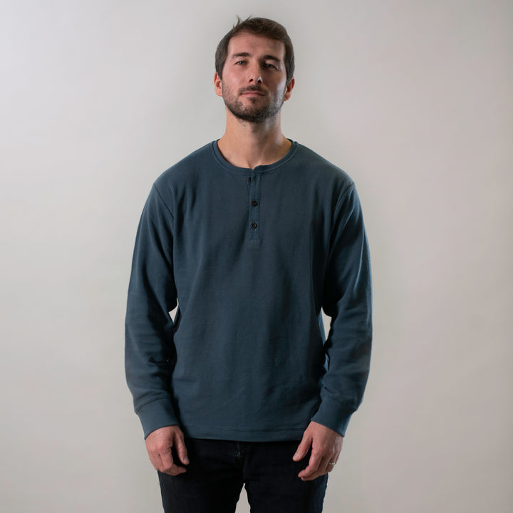 A man with short brown hair and a beard stands against a plain light background, wearing the Men's Elko Henley in Atlantic Blue and black pants, with a neutral expression and his hands relaxed by his sides.
