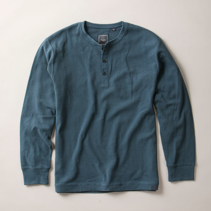 The Men's Elko Henley in Atlantic Blue, a cotton waffle classic fit shirt with long sleeves and three collar buttons, is laid flat on a light surface. It features a textured fabric and a simple, relaxed design.