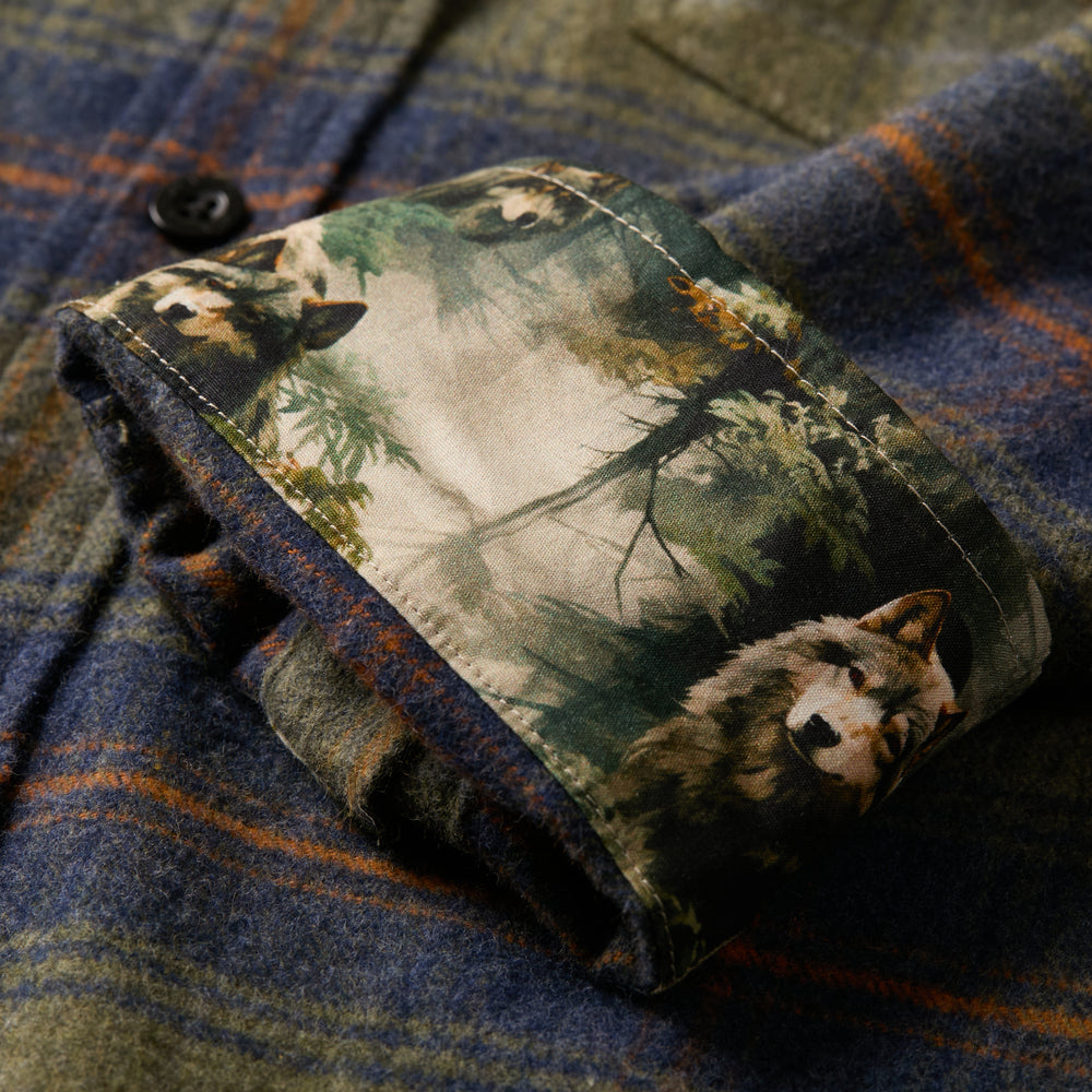 A close-up of the Men's Every Day Flannel Shirt in Everett Green Heather shows its olive, blue, and brown plaid pattern with the cuff folded back to reveal an inner lining adorned with wolves and trees.