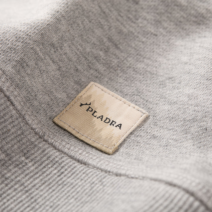 A close-up of the Men's Klamath Crew - Fog Grey premium cotton fabric shows a beige rectangular tag with the PLADRA brand name and a branch graphic—a detail from their classic fit sweatshirt.