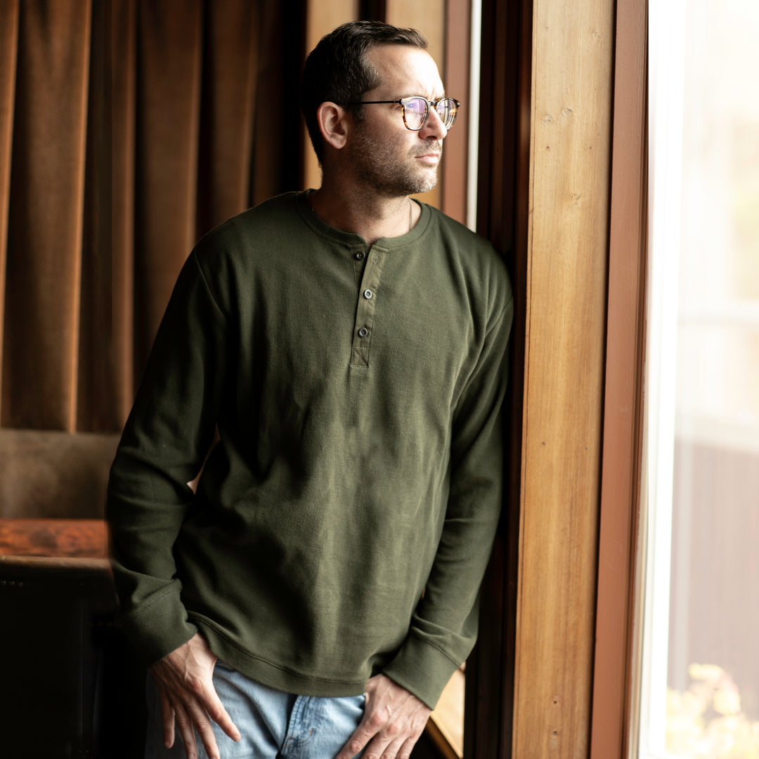 Man in glasses and forest green Pladra Elko Henley stands by a window, exuding thoughtful style.