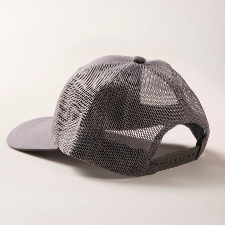 The Mountain Trucker Hat- Charcoal Grey is displayed from the back and side, featuring breathable mesh and an adjustable snapback strap against a light background.