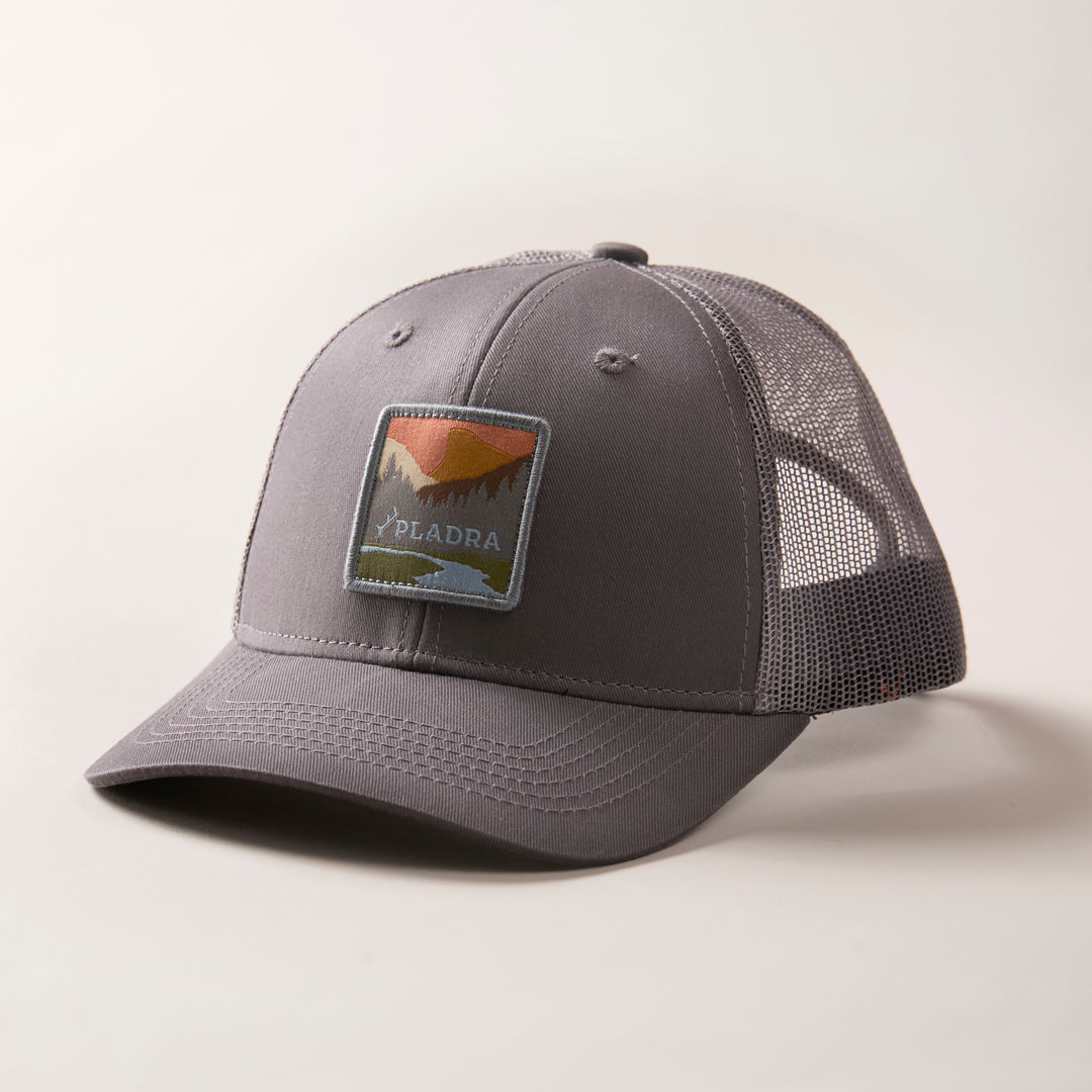 The Mountain Trucker Hat- Charcoal Grey features a mid-crown gray mesh design, a mountain landscape patch with "PIADRA" below, and an adjustable snap back for comfort. It is displayed on a plain, light-colored surface.