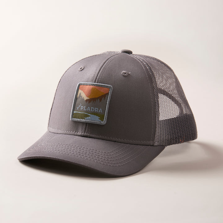 The Mountain Trucker Hat- Charcoal Grey features a mid-crown gray mesh design, a mountain landscape patch with "PIADRA" below, and an adjustable snap back for comfort. It is displayed on a plain, light-colored surface.