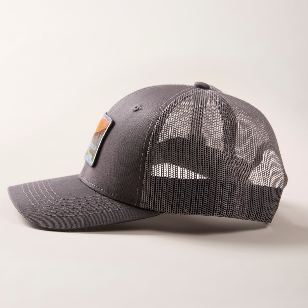 The Mountain Trucker Hat- Charcoal Grey features a curved brim, mesh back panels, and a colorful rectangular front patch. Displayed on a light background, it has an adjustable snap back for a comfortable fit.