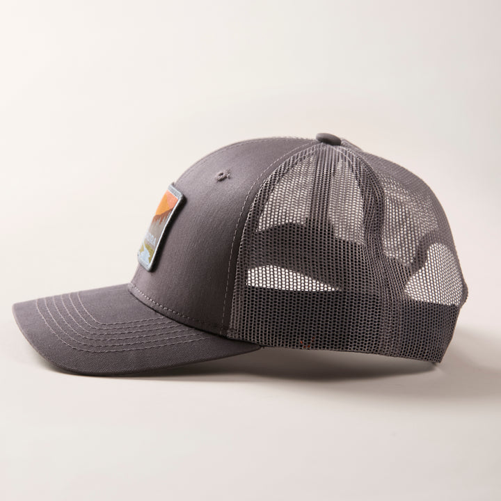 The Mountain Trucker Hat- Charcoal Grey features a curved brim, mesh back panels, and a colorful rectangular front patch. Displayed on a light background, it has an adjustable snap back for a comfortable fit.