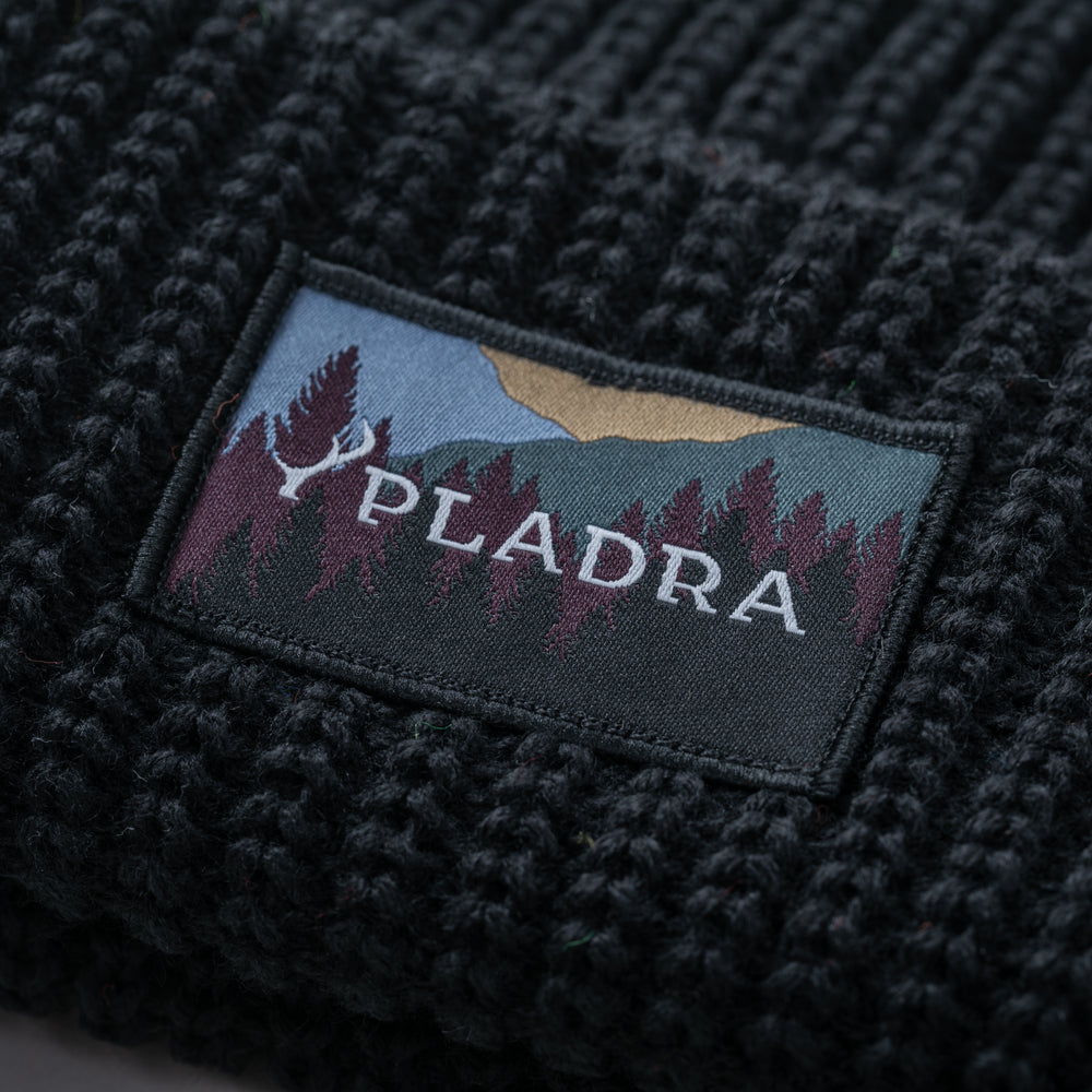 Close-up of the Woodsman Heavyweight Cuff Beanie in black with a Pladra patch featuring trees, mountains, and logo.