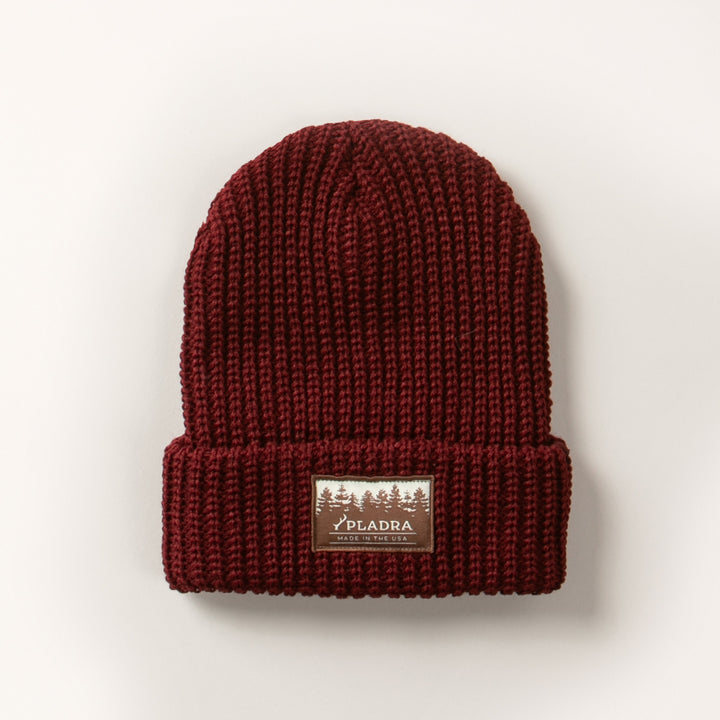The Woodsman Heavyweight Cuff Beanie in Burgundy Red is made in the USA, features a folded brim with a rectangular patch label of trees and PELADRA text on the front, and is shown on a plain white background.