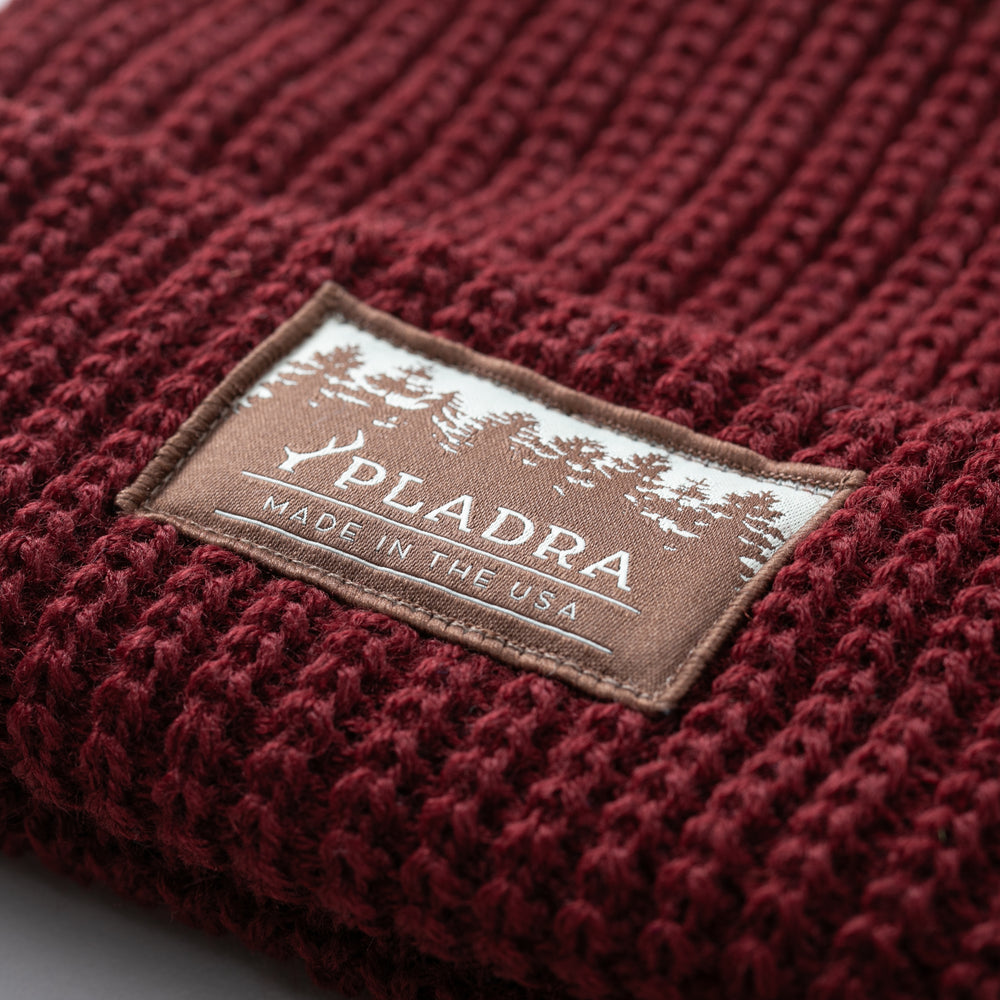 A close-up of the Woodsman Heavyweight Cuff Beanie in Burgundy Red, featuring a rectangular PLADRA MADE IN THE USA patch with tree silhouettes.