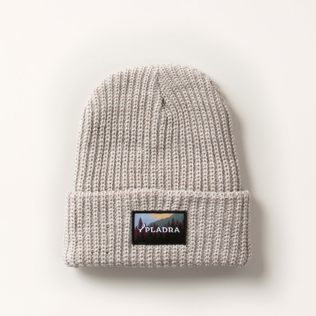 The Woodsman Heavyweight Cuff Beanie in Cloud White is a light gray knit with a folded brim and front rectangular patch showing a mountain landscape and "PLADRA." This beanie is made in the USA and shown on a plain white background.