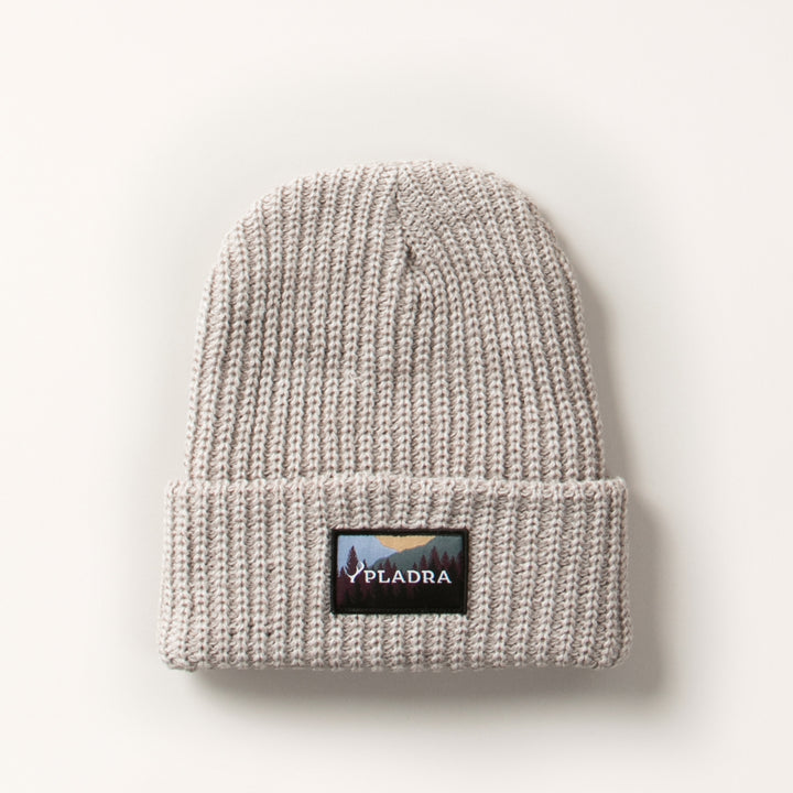 The Woodsman Heavyweight Cuff Beanie in Cloud White is a light gray knit with a folded brim and front rectangular patch showing a mountain landscape and "PLADRA." This beanie is made in the USA and shown on a plain white background.