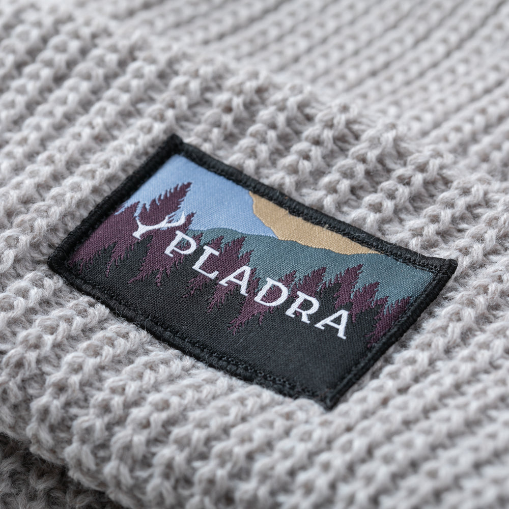 A close-up of the Woodsman Heavyweight Cuff Beanie- Cloud White, a light gray knit with a rectangular patch showing stylized trees, mountains, and "PLADRA" in white. Made in USA.
