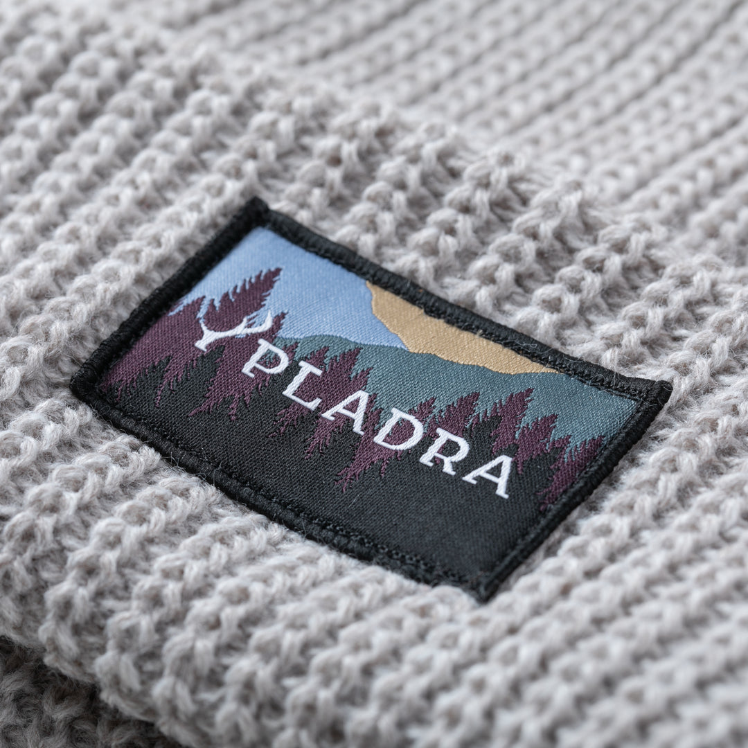 A close-up of the Woodsman Heavyweight Cuff Beanie- Cloud White, a light gray knit with a rectangular patch showing stylized trees, mountains, and "PLADRA" in white. Made in USA.