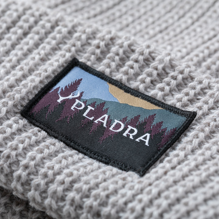 A close-up of the Woodsman Heavyweight Cuff Beanie- Cloud White, a light gray knit with a rectangular patch showing stylized trees, mountains, and "PLADRA" in white. Made in USA.
