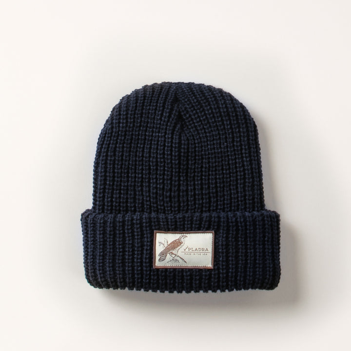 The Woodsman Heavyweight Cuff Beanie in Dark Navy Blue features a folded brim with a white rectangular patch displaying text and an illustration at the front center. Made in the USA.
