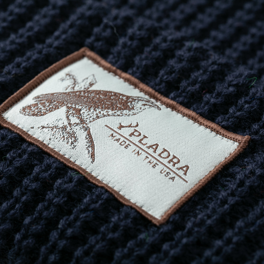 Close-up of a fabric label stitched onto dark navy blue knit material from the Woodsman Heavyweight Cuff Beanie—Dark Navy Blue. The label displays a bird illustration and brown text: PLADRA MADE IN THE USA.