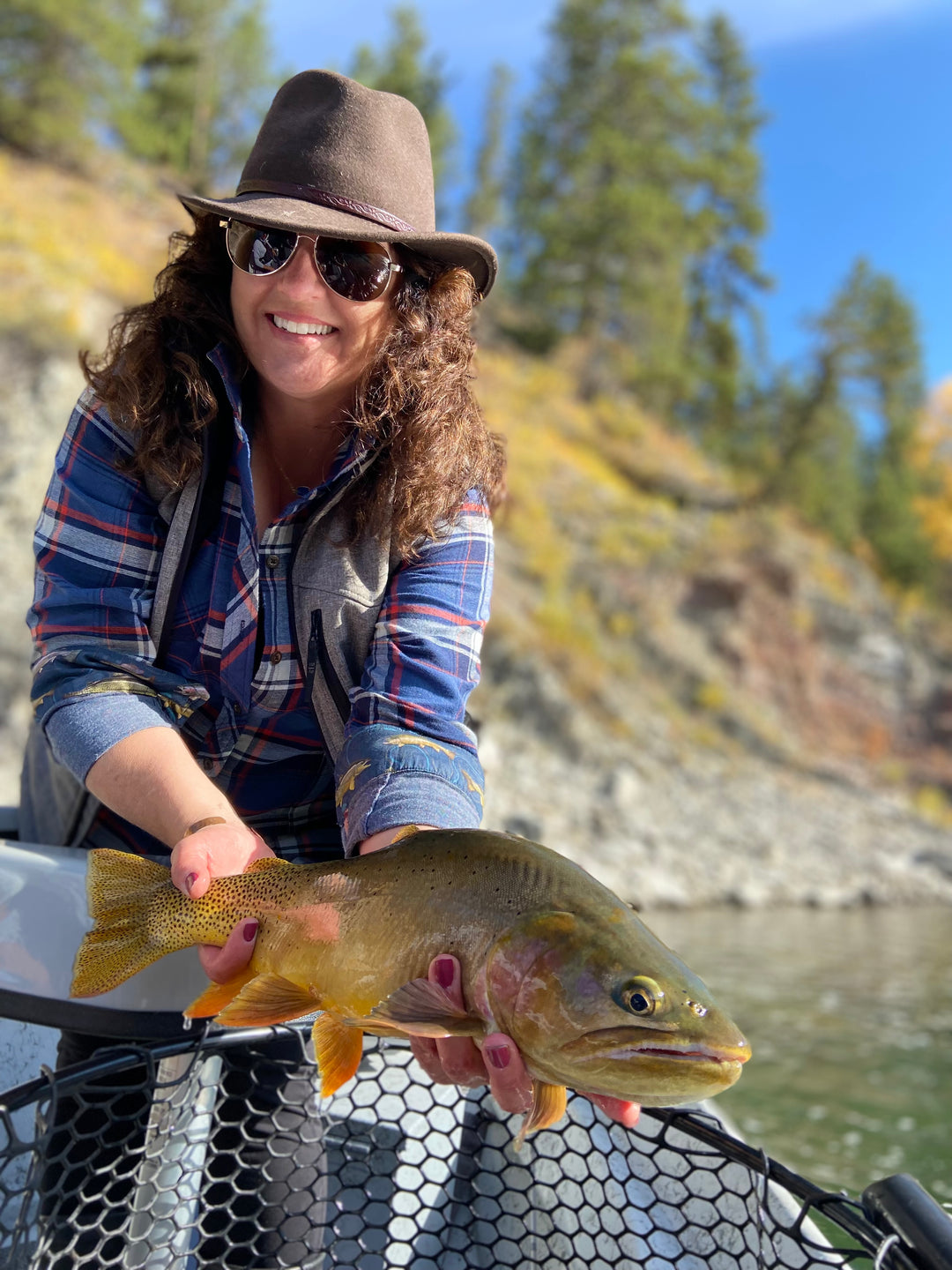 A smiling woman in sunglasses holds a large fish by a river, with trees and rocks in the background.