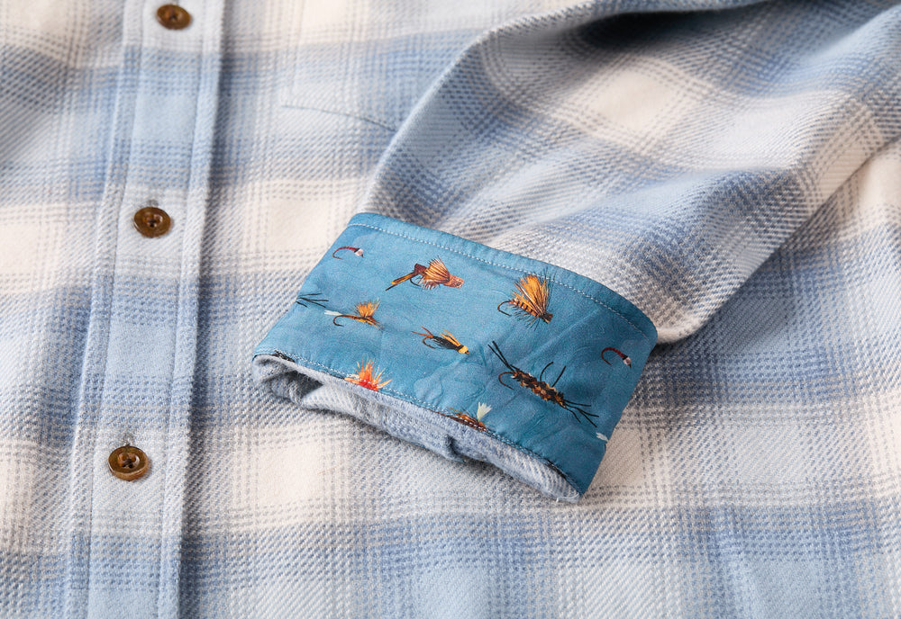 The Men's Every Day Elite Flannel Shirt in Mendocino Blue features a light blue plaid, brown buttons, and a cuffed sleeve showing an inner lining with colorful fishing lure designs—ideal as an outdoor flannel for cool days.