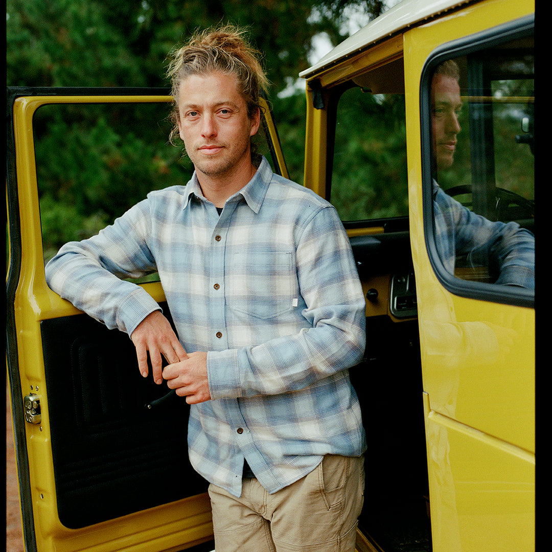 A person with blond hair in a messy bun stands by an open yellow van door, wearing the Men's Every Day Elite Flannel Shirt in Mendocino Blue and beige pants, with trees in the background.