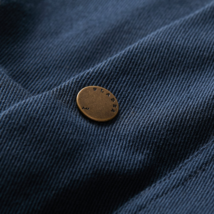 A close-up of a brass button embossed with PLADRA, attached to dark blue corduroy from the Men’s Sequoia Chore Coat in Midnight Blue.