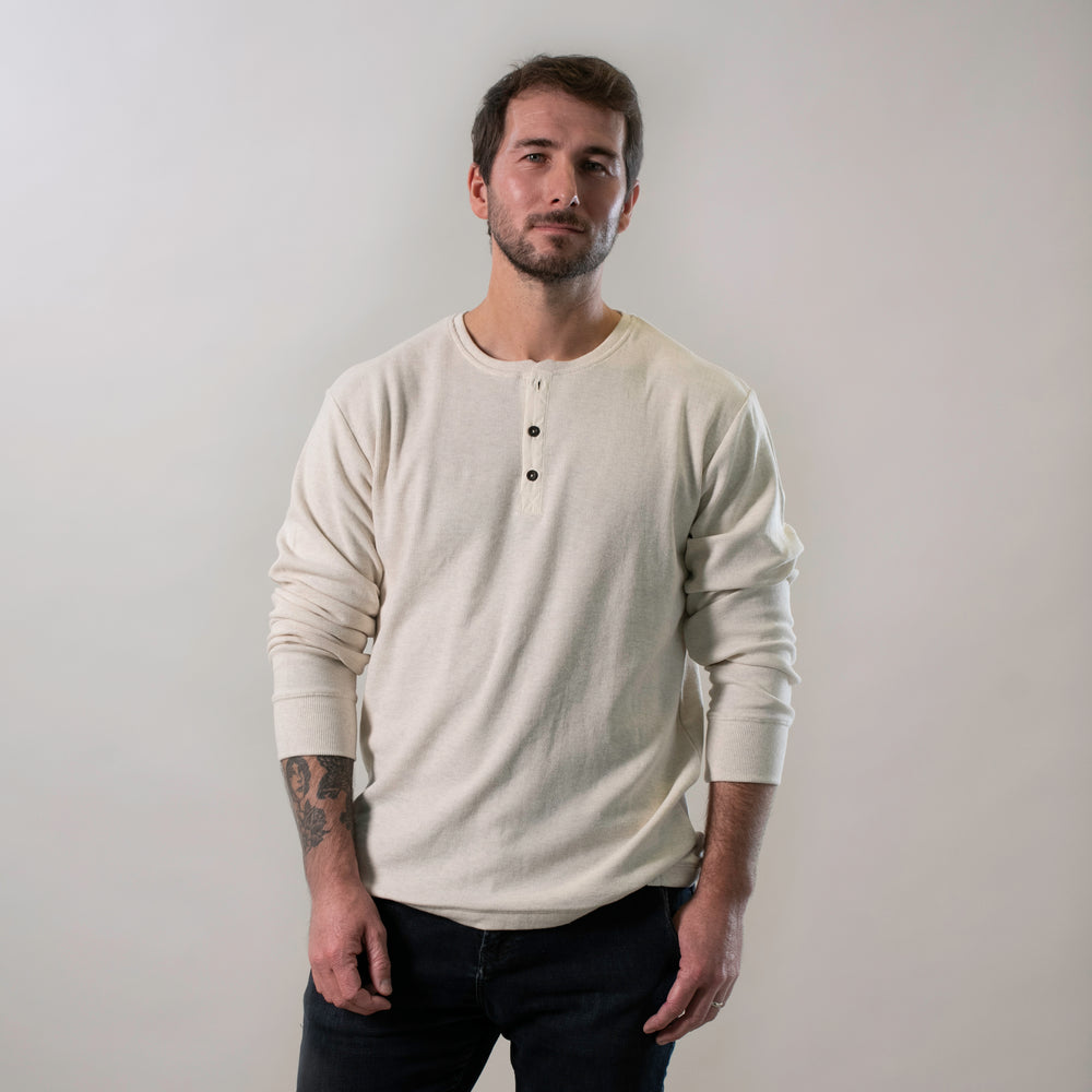 A man with short brown hair and a beard stands against a plain light background, wearing the Men's Elko Henley in Oatmeal White and dark jeans. He has a neutral expression, with one arm displaying a visible tattoo.